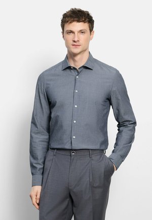 Mann trägt ein eng anliegendes, graues, langärmeliges Hemd mit Muster, das in graue Anzugshosen gesteckt ist, steht mit einer Hand in der Tasche.