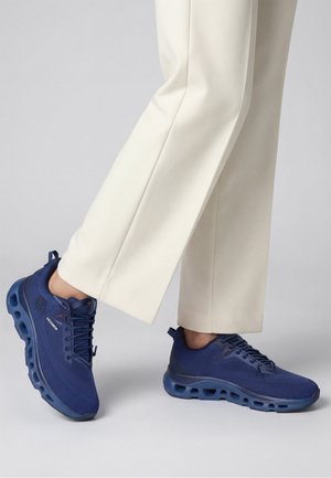 Chaussures de sport bleu marine avec semelles ventilées portées avec un pantalon couleur crème sur un fond gris clair.