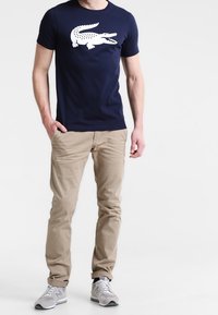 T-shirt en coton bleu marine avec un graphique de crocodile blanc, associé à un pantalon beige et des baskets grises. Ensemble simple et décontracté.