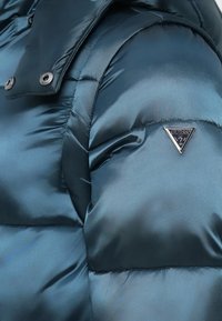 Manga de una chaqueta de plumas brillante y azul con el logo de Guess en forma de triángulo plateado y botones de metal en la tela acolchada cerca del hombro.