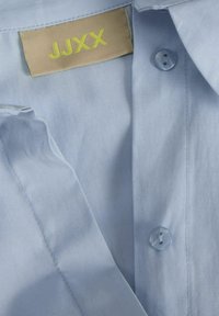 Lichtblauw katoenen overhemd met een kraag, voorzien van een gele label "JJXX" en een enkele knop bij de kraag. Gladde textuur en nette stiksels.
