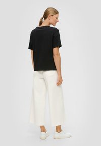 Zwarte T-shirt met korte mouwen en een horizontale ribbelstructuur, gecombineerd met wijd-uitlopende witte broeken met rafelige uiteinden en witte sneakers.