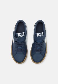 Jūras zila Nike sporta apavi ar gludu sintētisku virsu, baltām logo akcentiem, Velcro jostu un brūnu gumijas zoli.