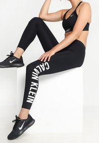 Czarny stanik sportowy, czarne legginsy z wysokim stanem z napisem "CALVIN KLEIN" po bokach oraz czarne buty sportowe o teksturowanym wykończeniu.