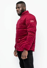 Giubbotto imbottito bordeaux con una texture liscia, chiusura con zip, polsini a costine e patch con marchio sulla parte superiore del braccio. Presenta due tasche frontali.