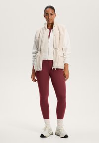 Witte fleece vest over een rode top en bijpassende leggings, met witte sneakers met een textured zool. Glad, aansluitend textiel en neutrale tinten.