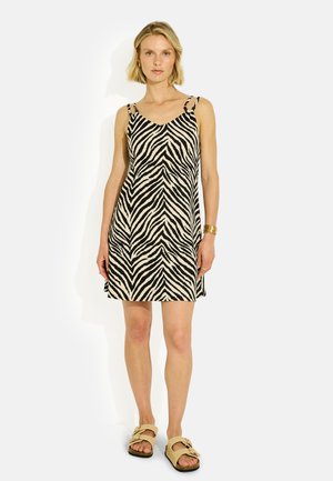 Mujer de pie con vestido sin mangas con estampado de cebra en negro y beige y sandalias beige sin talón con pulsera dorada en la muñeca izquierda.
