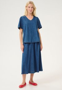 Denim divu daļu komplekts, kas sastāv no V-veida kakla blūzes ar īsām piedurknēm un saskaņota garā svārka. Tumši zilā krāsā ar nelielu tekstūru, kombinēts ar sarkaniem apaviem uz plakanā pacēluma.