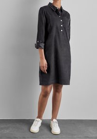 Abito camicia nero con colletto, chiusura con bottoni frontale, due tasche sul petto e maniche arrotolate, abbinato a sneakers bianche su uno sfondo grigio.