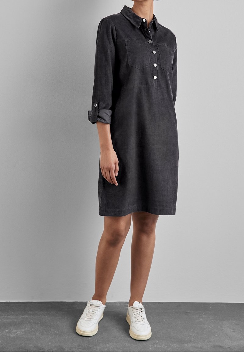 Abito camicia nero con colletto, chiusura con bottoni frontale, due tasche sul petto e maniche arrotolate, abbinato a sneakers bianche su uno sfondo grigio.