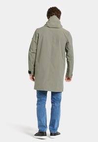 Hellgrüner wasserdichter Mantel mit hohem Kragen, langen Ärmeln und Rückenbelüftung. Kombiniert mit hellblauen Jeans und schwarzen Turnschuhen.