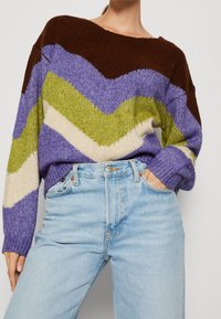 Person wearing a multicolored knit sweater with chevron stripes in brown, purple, green, and cream, paired with light blue high-waisted jeans.