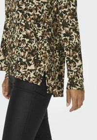 Langærmet bluse i beige med et mørkt blomstermønster, med sideslids og et tekstureret stof. Sammen med sorte tætsiddende bukser.