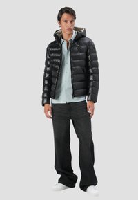 Giacca a vento nera con cappuccio, con zipper frontale e design trapuntato. Indossata sopra una camicia azzurra a bottoni e jeans scuri.