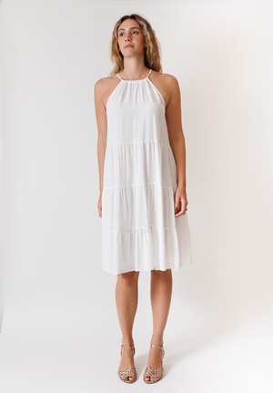 Abito bianco con scollo a halter e design a strati. Tessuto morbido, lunghezza al ginocchio, con spalline sottili e una silhouette fluida. Indossato con sandali con cinturini.