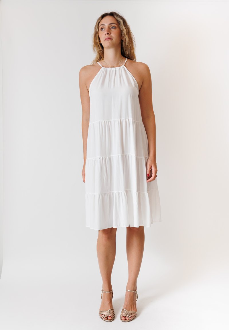 Abito bianco con scollo a halter e design a strati. Tessuto morbido, lunghezza al ginocchio, con spalline sottili e una silhouette fluida. Indossato con sandali con cinturini.
