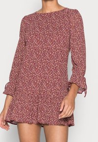 Frau in einem kastanienbraunen, blumigen Minikleid mit langen Ärmeln und Rüschenbündchen, die vor einem schlichten hellen Hintergrund steht.