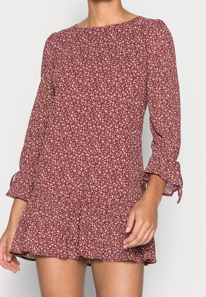 Frau in einem kastanienbraunen, blumigen Minikleid mit langen Ärmeln und Rüschenbündchen, die vor einem schlichten hellen Hintergrund steht.