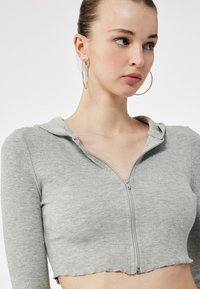 Grijze cropped zip-up hoodie gemaakt van ribbelstof, met een capuchon en een zilveren rits. Gladde textuur met ruwe zoomdetails.