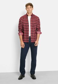 Rot-schwarzes kariertes Hemd mit umgeschlagenen Ärmeln, kombiniert mit navyfarbenen Hosen und schwarzen Schuhen. Das Modell steht vor einem schlichten Hintergrund.
