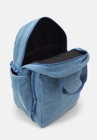 Mochila de denim en azul claro, con un gran compartimento principal, interior oscuro, asa superior y bolsillo con cremallera lateral. Textura suave.