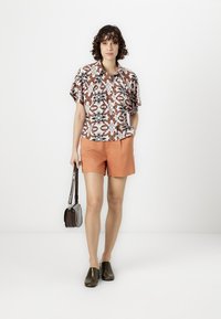 Short-sleeved, patterned blouse in white, orange, and blue; orange shorts; dark, shiny slip-on shoes; small brown handbag.