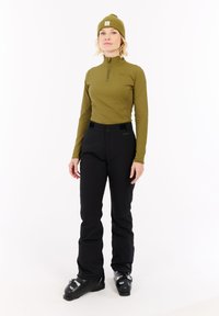 Vrouw in een olijfgroene longsleeve en beanie, zwarte skibroek en zwarte skischoenen staat tegen een witte achtergrond.
