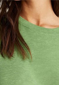 Gros plan sur le cou et l'épaule d'une personne portant un haut en tissu vert clair avec de longs cheveux bruns tombant sur le côté gauche.