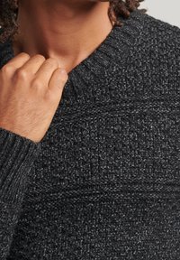 Dunkelgrauer Strickpullover mit strukturiertem Muster, geripptem Kragen und Bündchen sowie grober Webart. Der Stoff wirkt weich und warm.