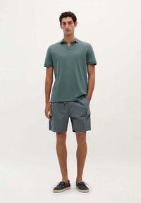 Grøn polo skjorte med krave, parret med grå cargo shorts med sidelommer. Modellen har marineblå sko med en lys sål på.