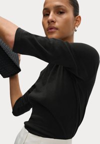 Svart, figurnära topp med trekvartslånga ärmar, mjukt material och en lätt lyster, kombinerad med en vävd svart handväska. Minimalistisk design.