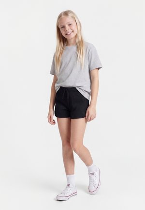 Uśmiechnięta dziewczyna z długimi blond włosami, ubrana w szary t-shirt, czarne szorty, białe skarpetki i białe trampki, stoi na białym tle.