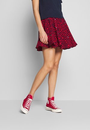 Kvinde iført et rødt nederdel med hjerteprint, marineblå top og røde sneakers med højt skaft, stående med krydsede ben mod en grå baggrund.