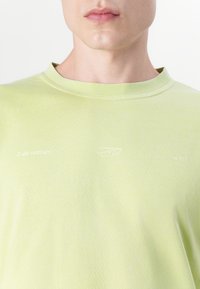 Light green T-shirt with a crew neckline, featuring subtle texture and embroidered branding on the chest and sleeves.