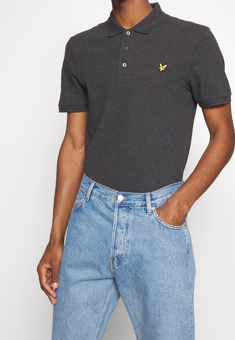 Homme portant un polo à manches courtes gris foncé avec un logo oiseau jaune, rentré dans un jean taille haute bleu clair, une main dans la poche.