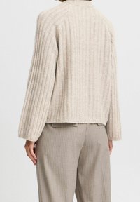 Suéter de punto acanalado en color beige con un cuello alto, hombros caídos y un dobladillo ligeramente recortado, combinado con pantalones de rayas finas en gris claro.