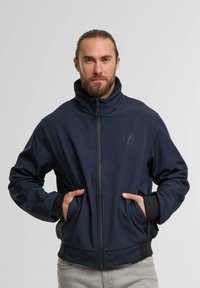 Marineblaue Zip-Jacke mit hohem Kragen, Fronttaschen und schwarzen Akzenten an den Bündchen und dem Saum. Weicher, glatter Stoff mit minimalistischer Logoprägung.
