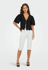 Crni kratki top s pripijenim rukavima i dubokim V-izrezom, s valovitim rubom i rupičastim detaljima, uparen s bijelim kratkim hlačama.