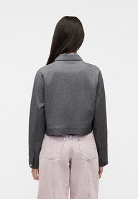 Γυναίκα με μακριά σκούρα μαλλιά, φορώντας μια κοντή σκούρα γκρι ζακέτα και ανοιχτό μωβ ψηλόμεσα τζιν, με την πλάτη προς τον θεατή.