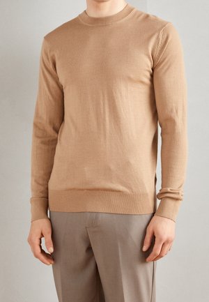 Mand iført en tætsiddende kamelfarvet langærmet sweater og lysegrå bukser, der står mod en ensfarvet baggrund.