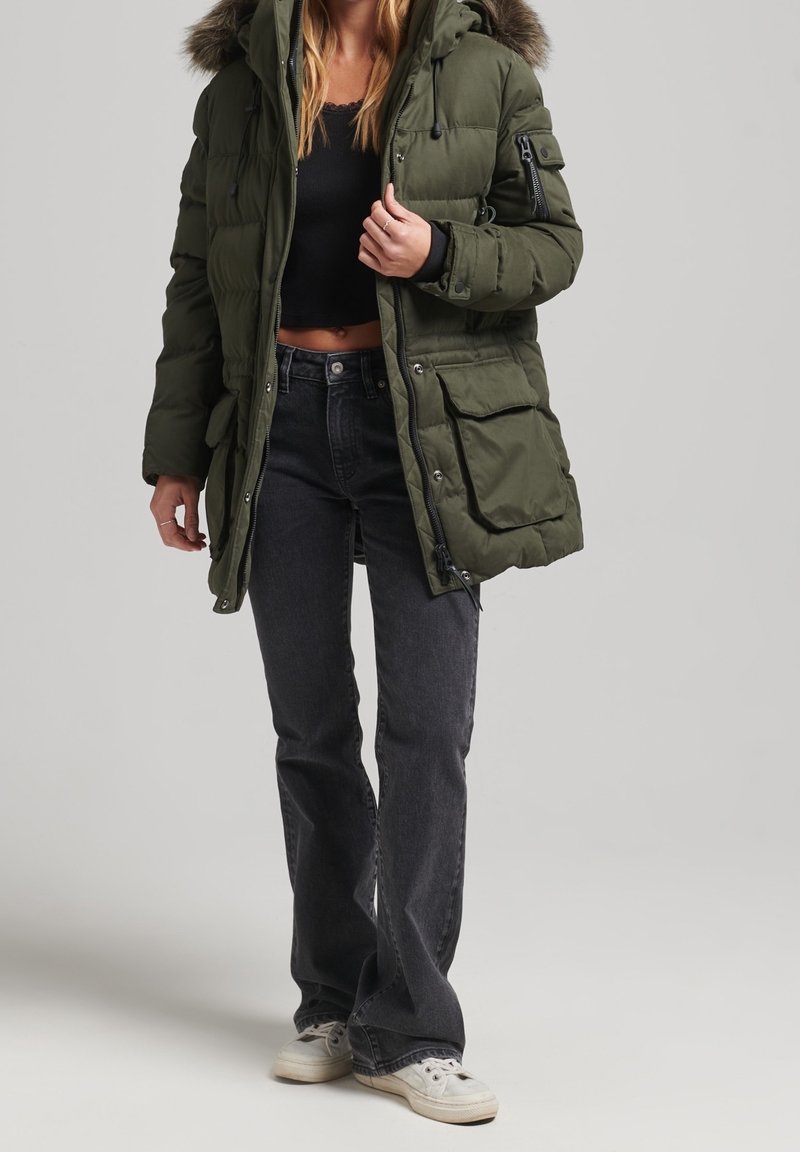 Femme portant une parka matelassée vert olive avec capuche en fourrure, un crop top noir, un jean évasé foncé et des baskets blanches, debout sur un fond clair.