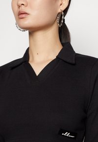 Top preto com textura canelada e decote em V, apresentando uma pequena etiqueta preta com texto branco. Grandes brincos de argola prateados torcidos também são visíveis.