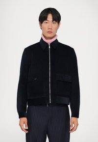 Navy corduroy jacket with a zip front, two chest pockets, and a short, straight cut. Worn over a pink high-neck top.