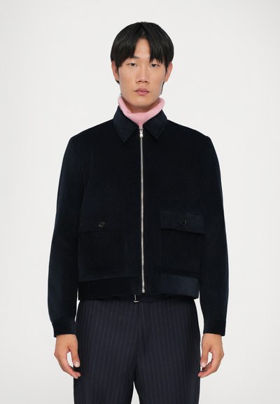 Veste en velours côtelé bleu marine avec fermeture éclair sur le devant, deux poches poitrine et une coupe courte et droite. Portée par-dessus un top rose à col montant.