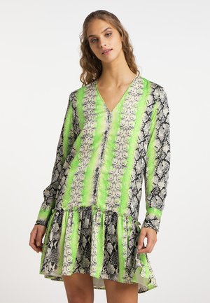 Młoda kobieta ubrana w sukienkę z długim rękawem i dekoltem w serek z neonowo zielonym i czarnym wzorem węża, stojąca na białym tle.