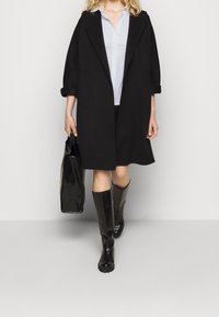 Cappotto oversize nero, camicia azzurro chiaro, stivali neri al ginocchio e borsa nera. Tessuto morbido, design minimalista e vestibilità ampia.