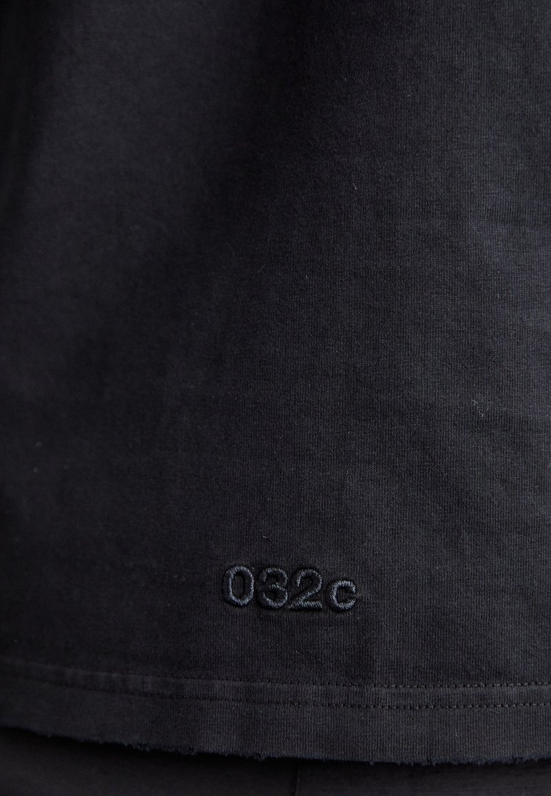 Schwarzes Baumwoll-T-Shirt mit kleinem, in passendem Schwarz gesticktem "032c"-Logo nahe dem Saum. Weicher Stoff mit klassischem Schnitt.