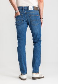 Mężczyzna ubrany w niebieskie dżinsy i białe trampki, stojący z jedną ręką w tylnej kieszeni, na tle jasnego, jednolitego tła.