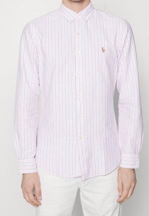 Uomo che indossa una camicia bianca a maniche lunghe con sottili righe verticali viola e verdi e un piccolo logo ricamato sul petto sinistro.