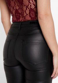 Black faux leather pants with a smooth texture, high waist, and stitched back pockets. The design features a form-fitting silhouette.